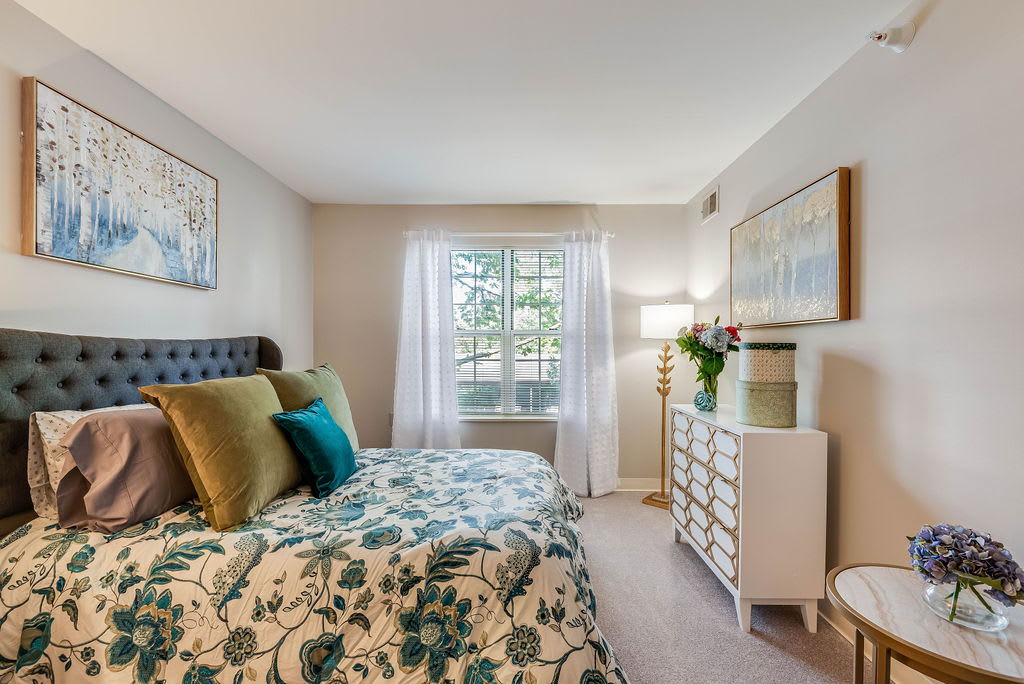 A cozy and well-decorated bedroom with patterned bedding, a cushioned headboard, and a bright window with sheer curtains. A dresser topped with fresh flowers stands next to a stylish floor lamp.