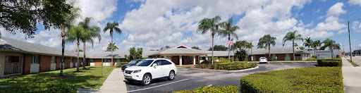 A modern building with palm trees in the foreground and a white car parked in the lot under a cloudy sky.