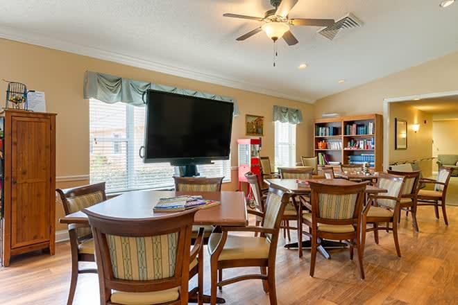 A cozy lounge area with a television, multiple chairs and tables, bookshelves, and large windows letting in natural light.