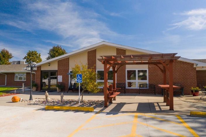 A small building with an entrance, flanked by a wooden pergola and surrounded by a parking area under a clear blue sky.