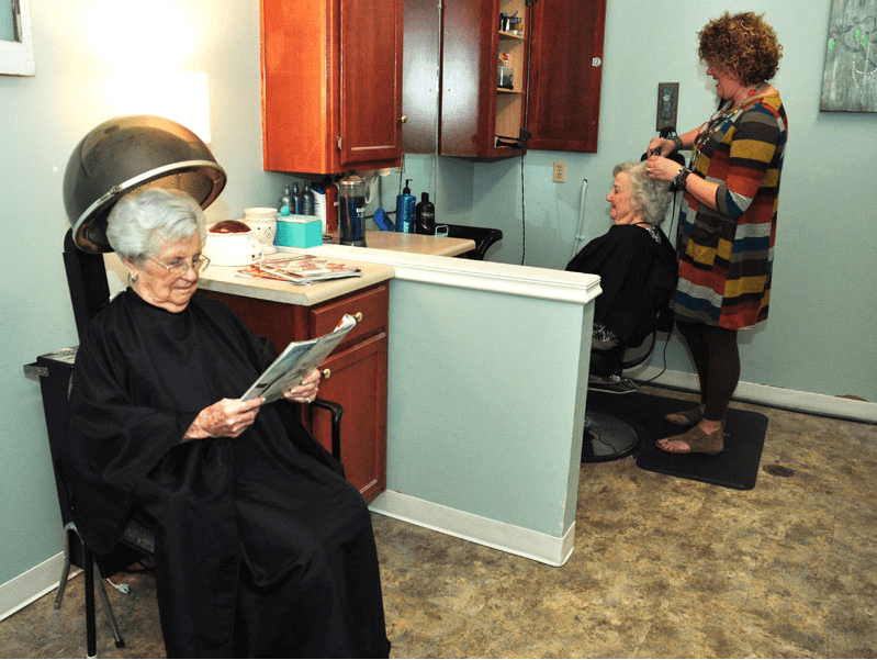 An elderly woman reads a magazine while having her hair dried in a salon, as another woman receives a haircut nearby.