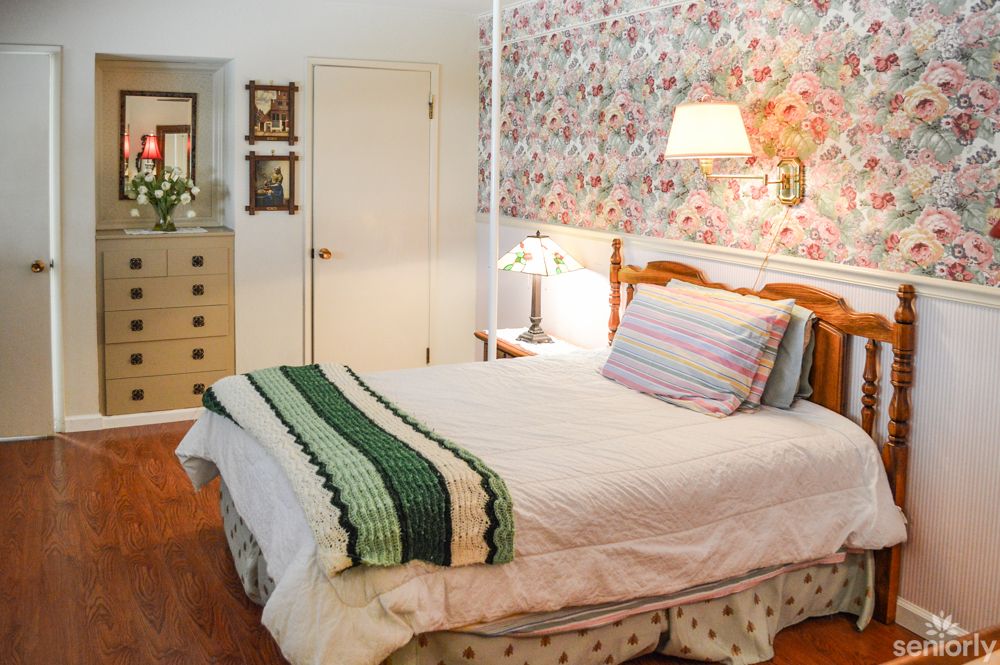 A cozy bedroom featuring floral wallpaper, a neatly made bed with a striped pillow and knitted blanket, and a wall-mounted lamp.