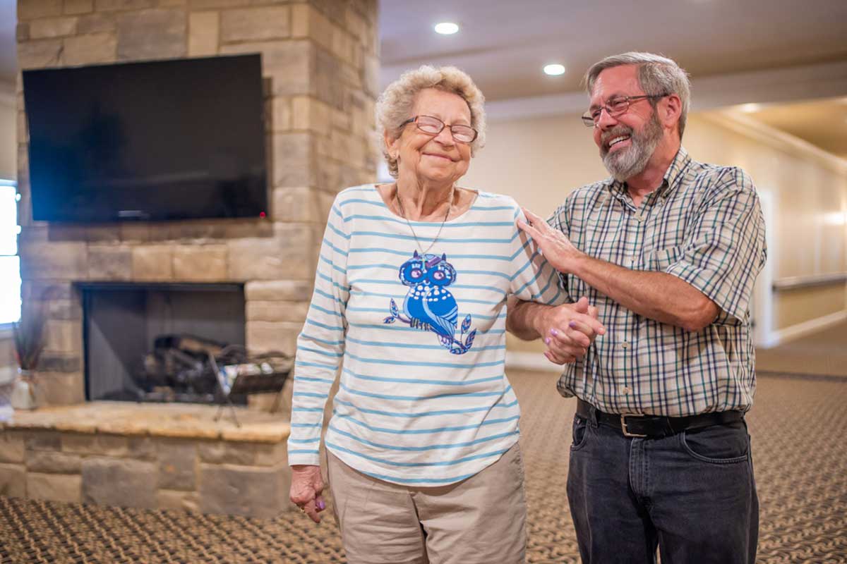 An elderly woman receiving support from a man indoors, both smiling happily.