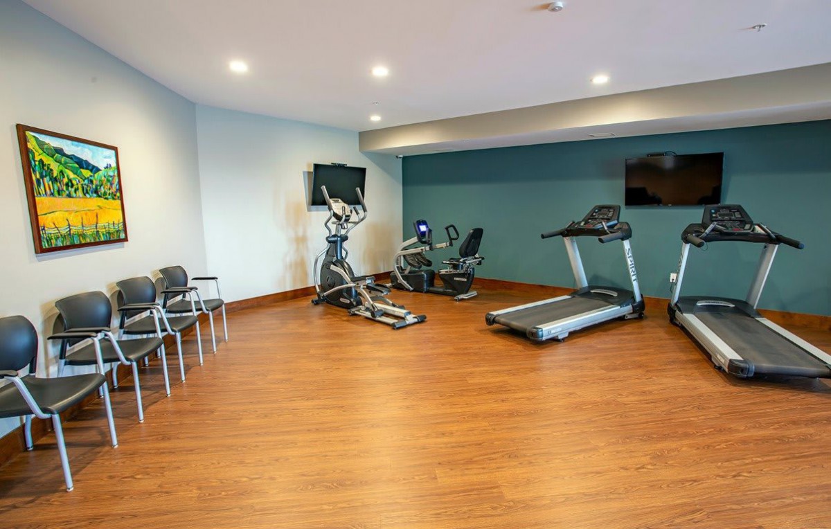 A small gym with exercise equipment including a treadmill, exercise bike, and elliptical machine on a wooden floor.