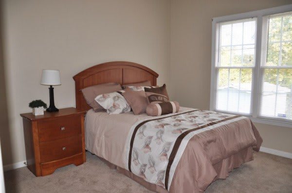 A neatly arranged bedroom with a bed featuring brown and beige bedding, a wooden headboard, a nightstand with a lamp, and a window with white trim.