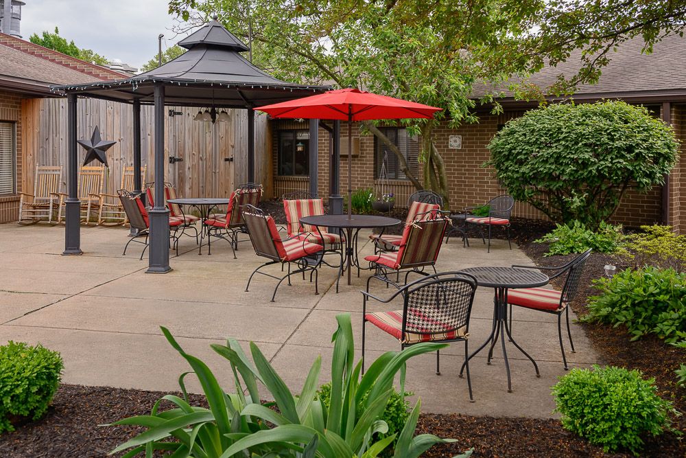 A cozy outdoor patio area featuring tables, chairs, a gazebo, and a red umbrella surrounded by lush greenery.