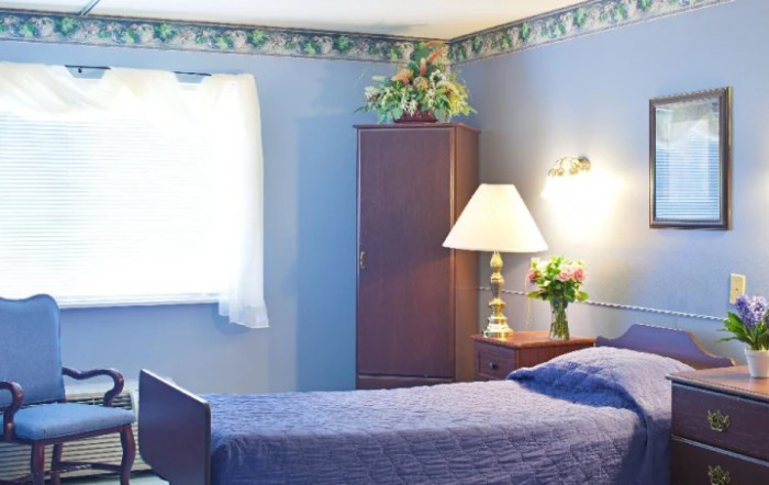 A cozy bedroom with blue walls, featuring a bed, a bedside table with a lamp, and a wardrobe.