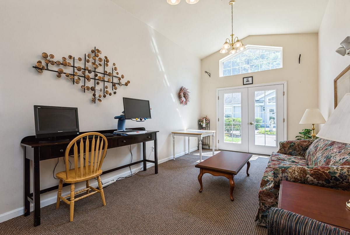 A cozy living room with a computer desk, wooden chair, coffee table, and sofa, illuminated by natural light through large windows.