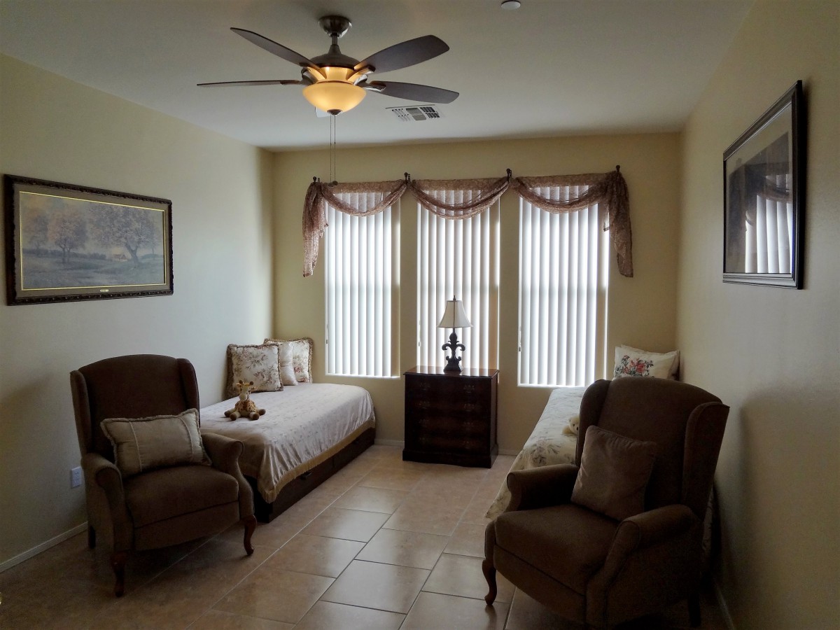 A cozy bedroom with two twin beds, chairs, and a ceiling fan, brightened by natural light streaming through tall windows adorned with curtains.