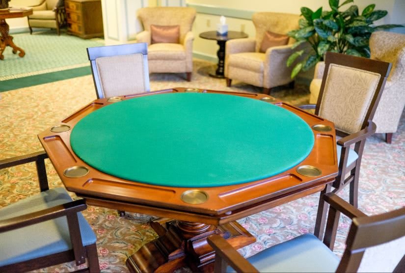 A wooden poker table with a green felt top surrounded by chairs in a cozy lounge area.