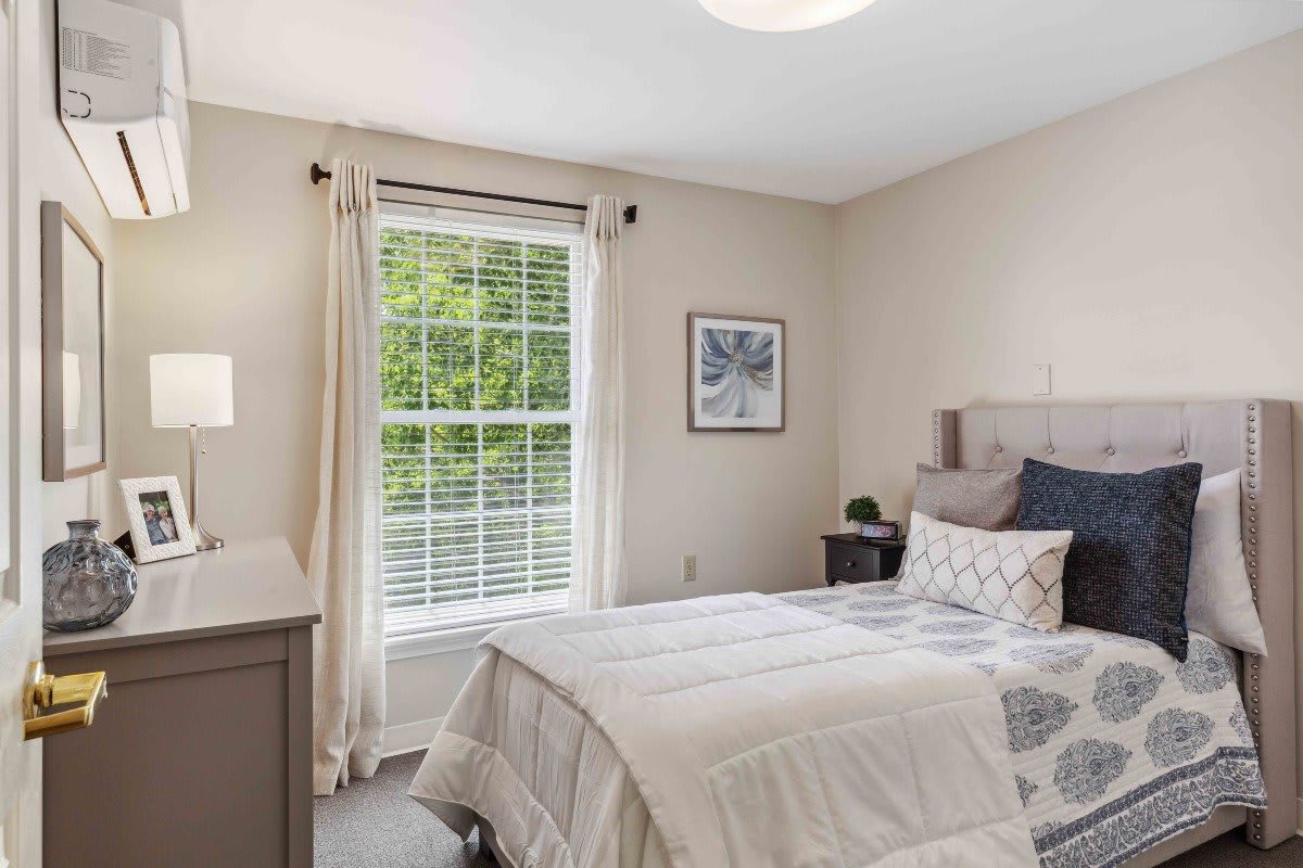 A cozy and well-lit bedroom featuring neutral tones, with a bed, lamp, window with curtains, and decorative artwork.