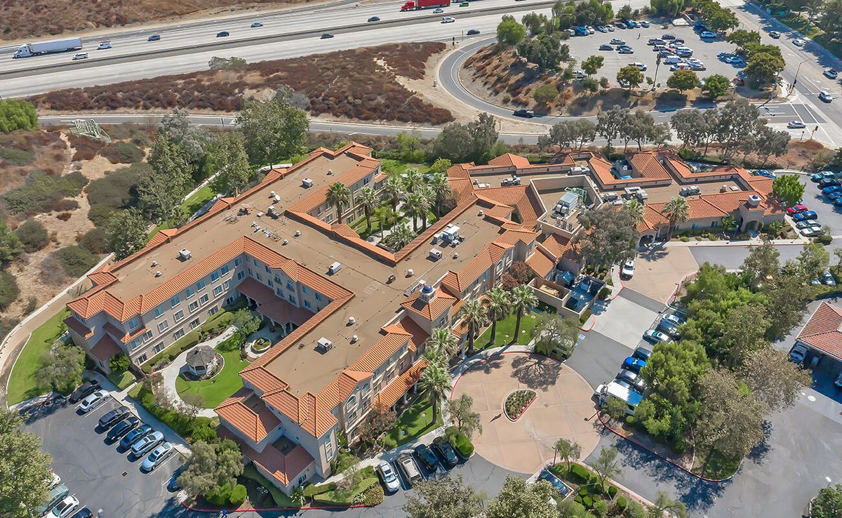 Aerial view of a large building complex with a parking lot and surrounding trees, near a highway.