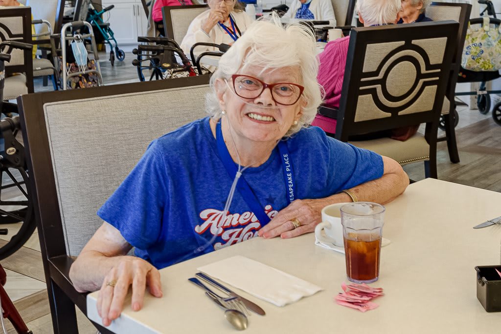 An elderly woman with glasses enjoying a meal at a restaurant, smiling warmly with a glass of iced tea in front of her.