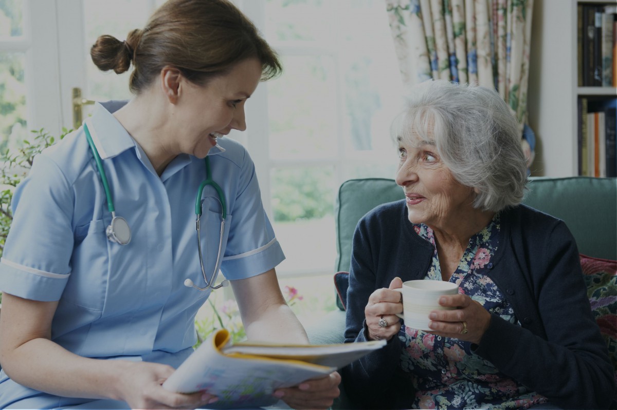 A nurse engages in conversation with an elderly woman who is enjoying a cup of tea.