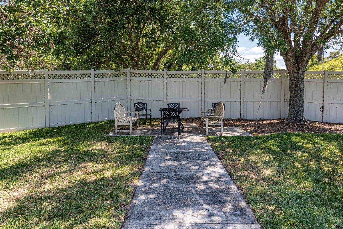 A backyard patio area with chairs and a table, surrounded by a white fence.