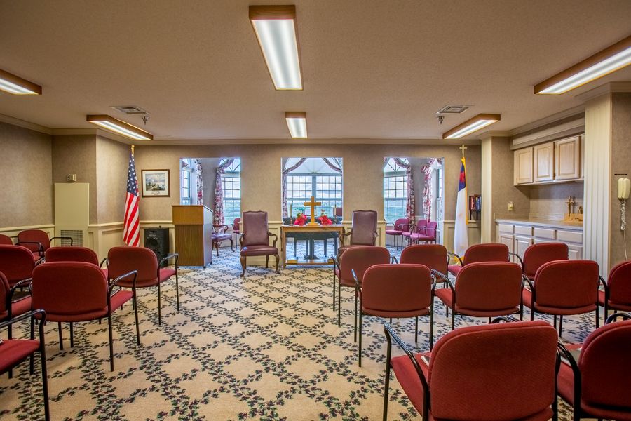 An indoor ceremony room with arranged chairs, decorative flooring, and flags displayed by the windows.