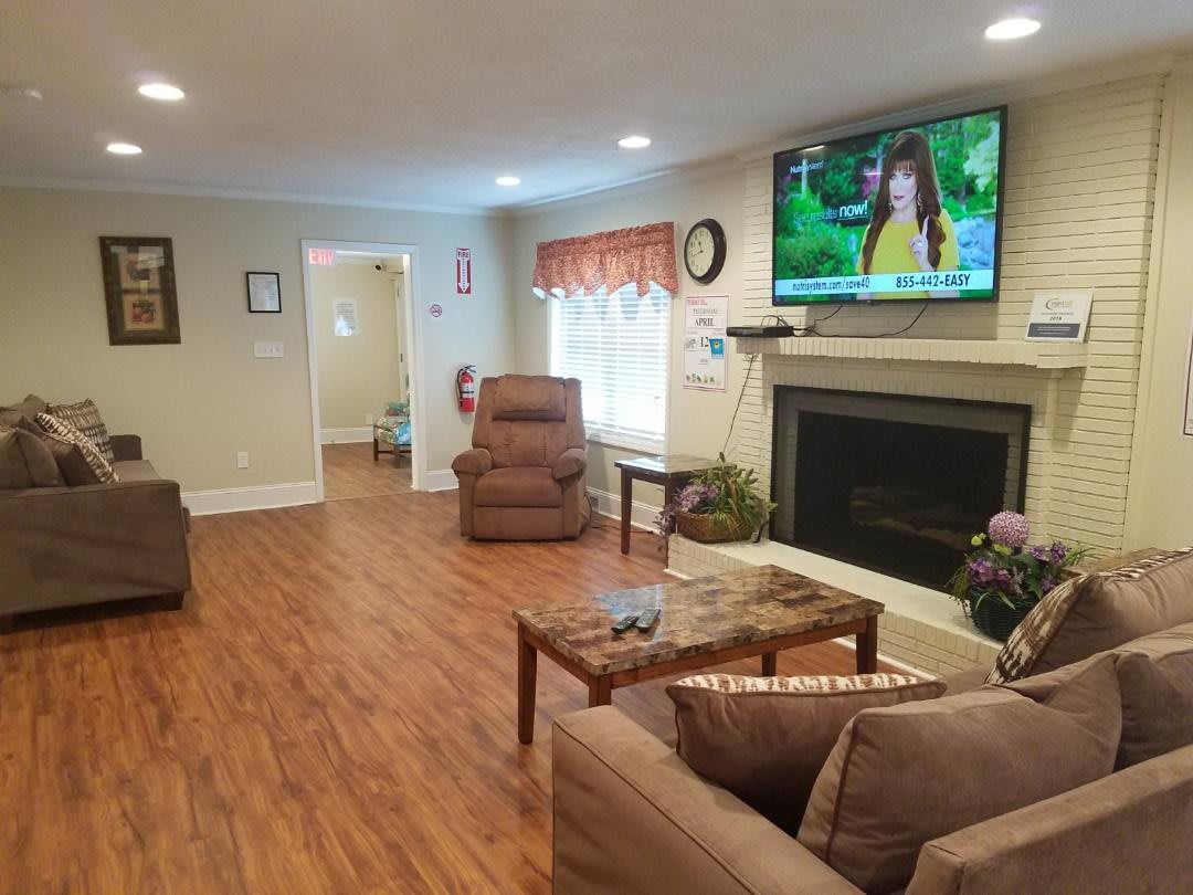 A cozy living room featuring a television above a fireplace, with recliner chairs and a sofa on wood flooring.