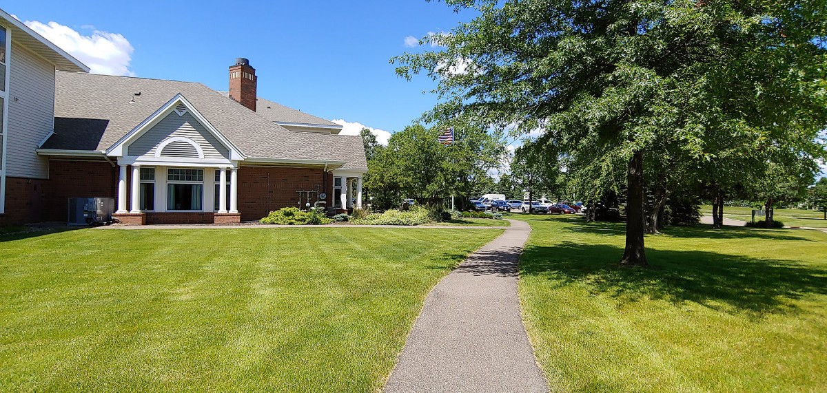 A sunny suburban scene with a brick house, well-maintained lawn, and a winding pathway lined by trees.