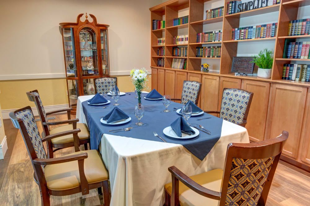 An elegantly set dining table surrounded by wooden bookshelves, featuring a blue and white tablecloth with folded napkins and a floral centerpiece.
