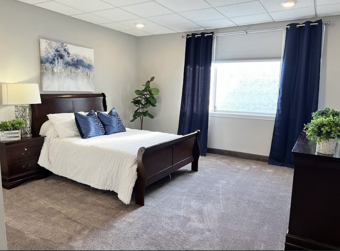 A well-decorated bedroom with a wooden bed frame, white bedding, and blue accent pillows. A large window with dark curtains allows natural light to fill the space.