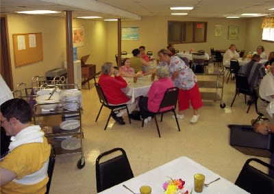 A group of seniors gather around a table in a dining area of a care facility.