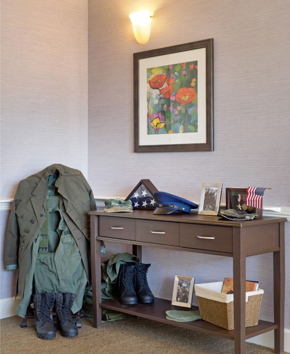 A military uniform and boots are neatly placed next to a table displaying a folded flag, medals, and family photos.
