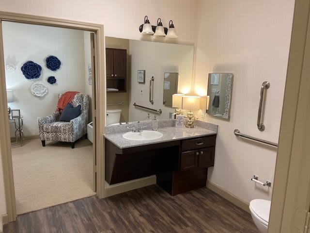 A spacious and accessible bathroom with a sink, mirror, lamp, and grab bars. The room features wood-style flooring and is connected to a cozy sitting area.