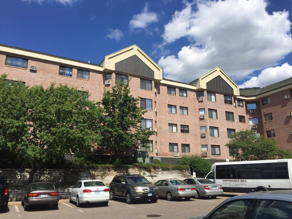 A view of a multi-story apartment building with a parking lot full of cars under a partly cloudy sky.