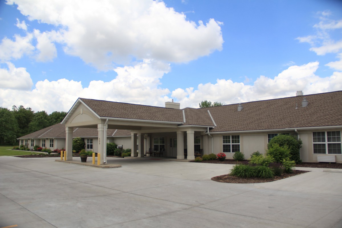 A one-story building with a covered entrance and driveway, set against a backdrop of clouds and blue sky.