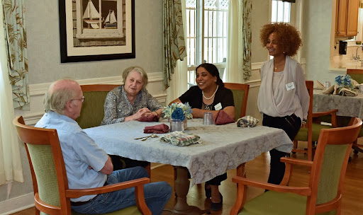 A group of elderly individuals engaging in conversation at a communal dining table with support staff.