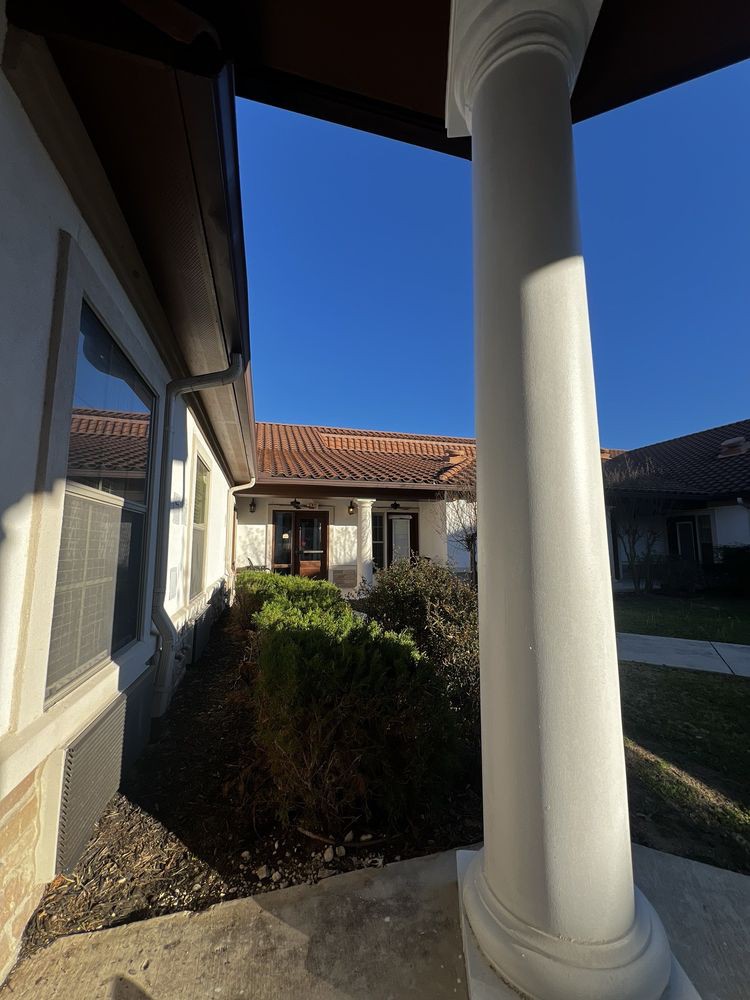 A sunny courtyard with a tiled roof, surrounded by bushes and white columns.