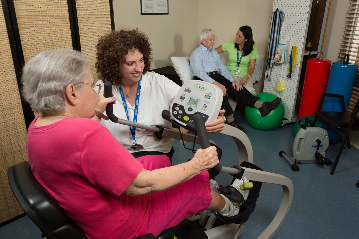 A healthcare professional assists a senior woman with exercise equipment in a therapy session, while another pair engages in conversation in the background.