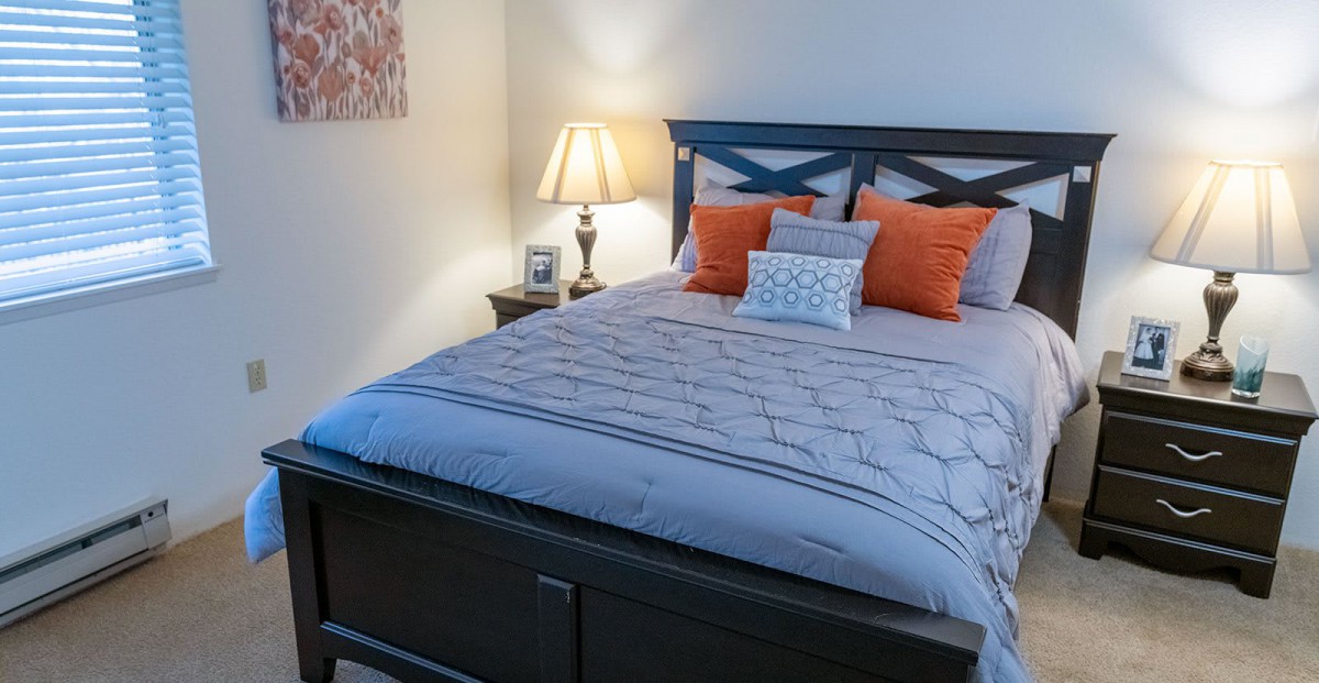 A cozy bedroom with a neatly made bed and colorful pillows, illuminated by a bedside lamp.