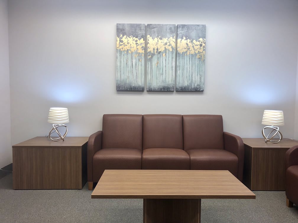 A modern office waiting area featuring a brown sofa, wooden table, lamps, and abstract artwork on the wall.