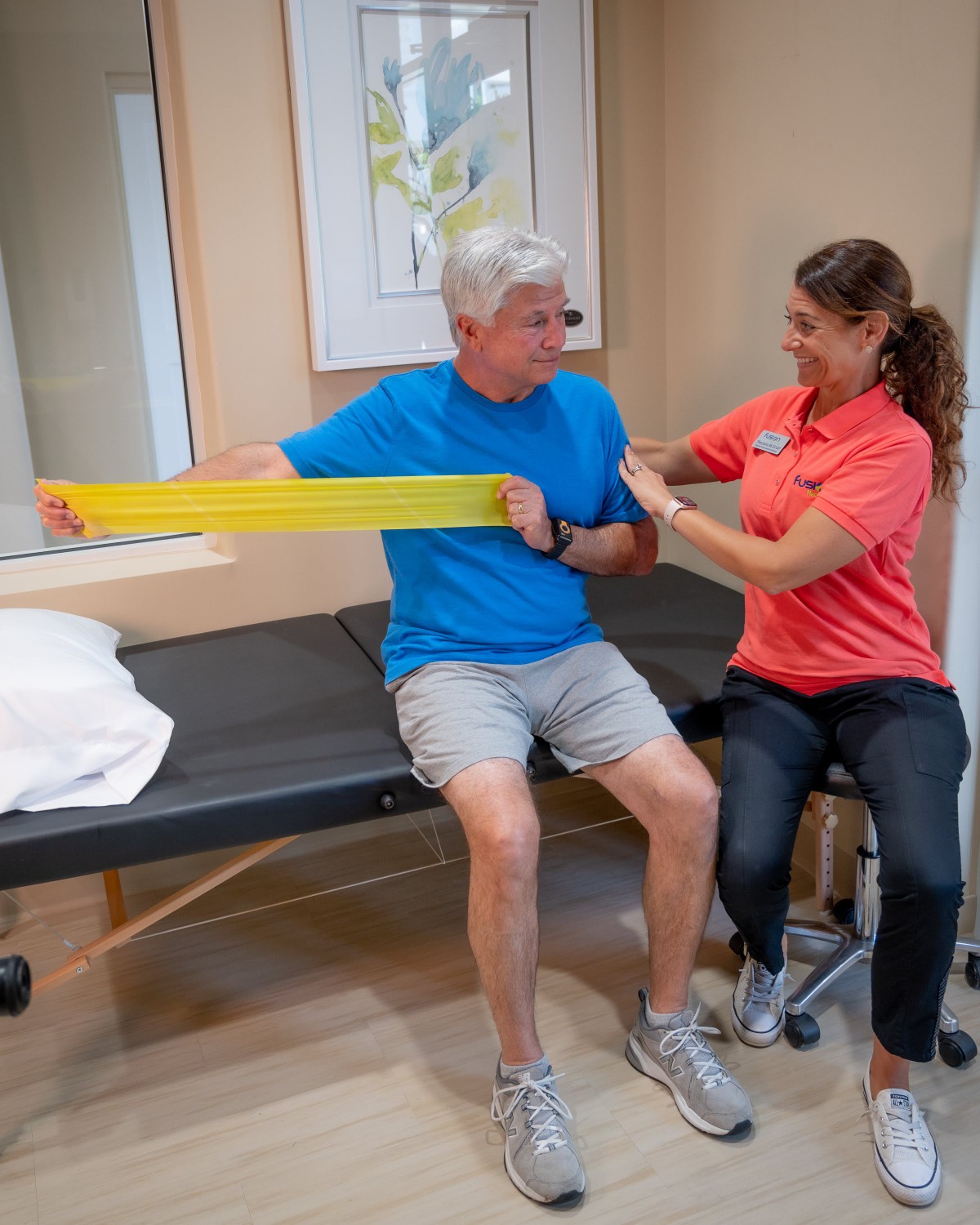 A senior man performs an exercise with a resistance band under the guidance of a therapist in a clinic setting.