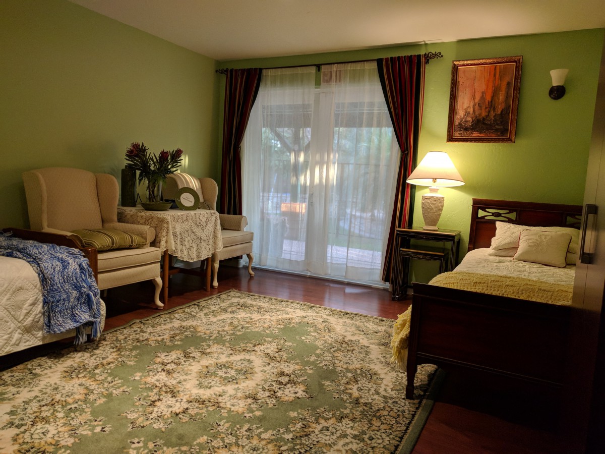 A cozy bedroom with traditional decor, featuring green walls, a patterned area rug, and elegant armchairs beside a large window with curtains.