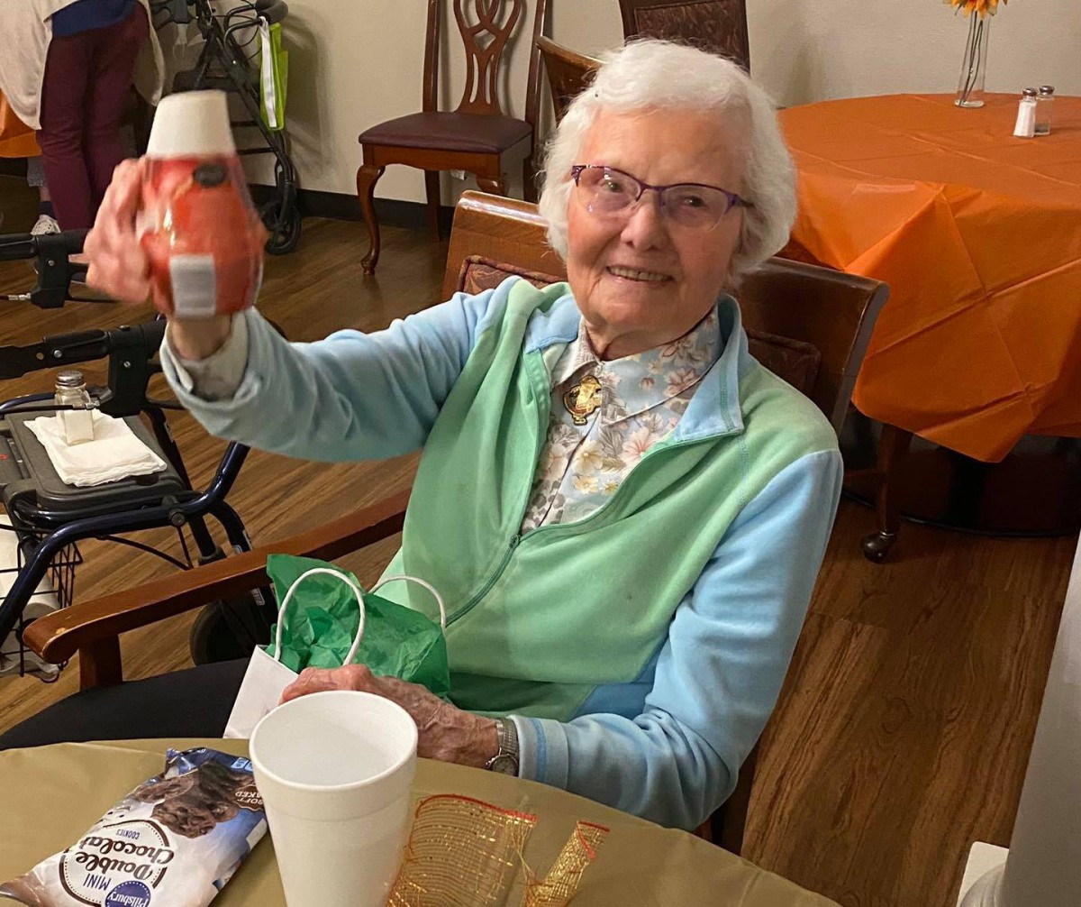 An elderly woman smiling and holding a bottle while sitting indoors.