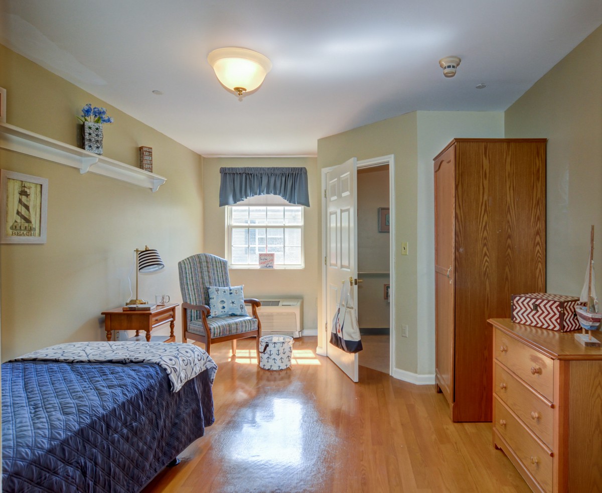 A cozy bedroom with a single bed, wooden furniture, and natural light from a window.