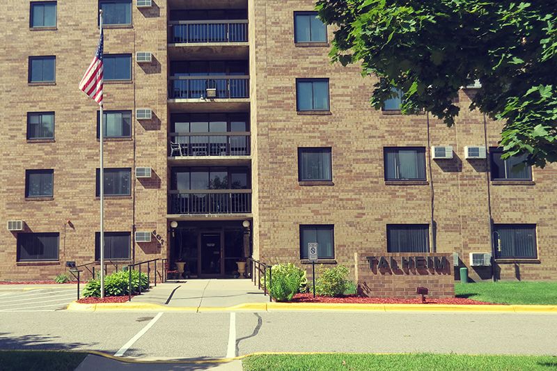 A brick apartment building with an entrance, surrounded by greenery and a flagpole in an urban setting.