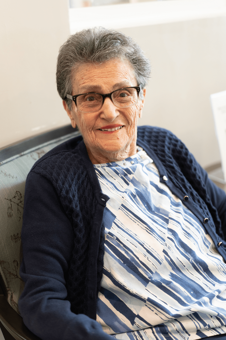 An elderly woman with glasses smiling warmly while seated indoors.