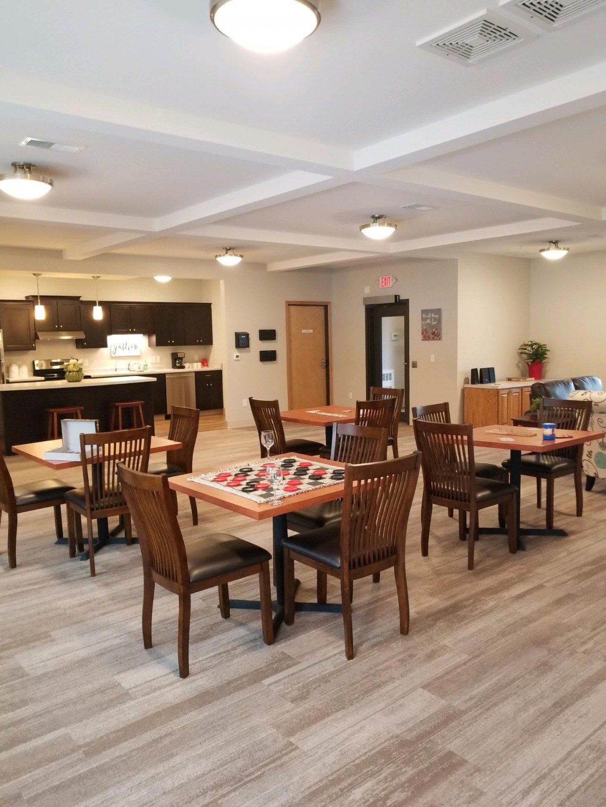 A spacious dining and community area with wooden tables and chairs, connected to a kitchen.
