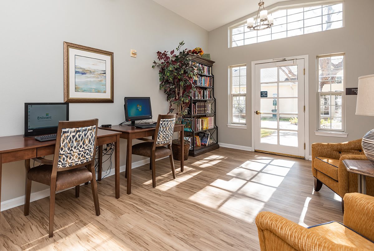 A bright home office featuring a desk with a computer, a bookshelf filled with books, and sunlight streaming through large windows.