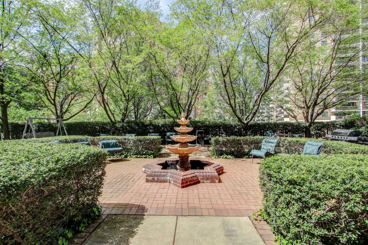 A tranquil garden with a central tiered fountain surrounded by lush trees and benches.