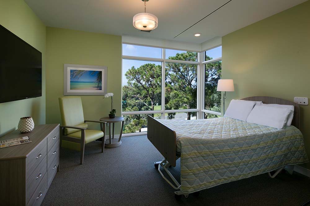 A hospital room with a large window offering a view of greenery outside, featuring a single bed, chair, and modern interior decor.