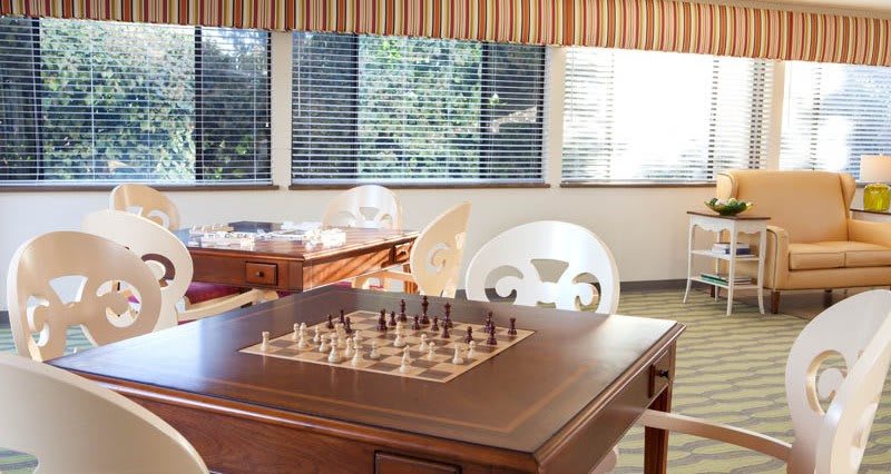 A cozy game room with a chessboard set up on a wooden table, illuminated by sunlight streaming through the windows.
