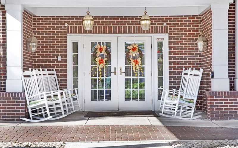 A welcoming entrance with double doors adorned with wreaths, flanked by white rocking chairs on a brick porch.