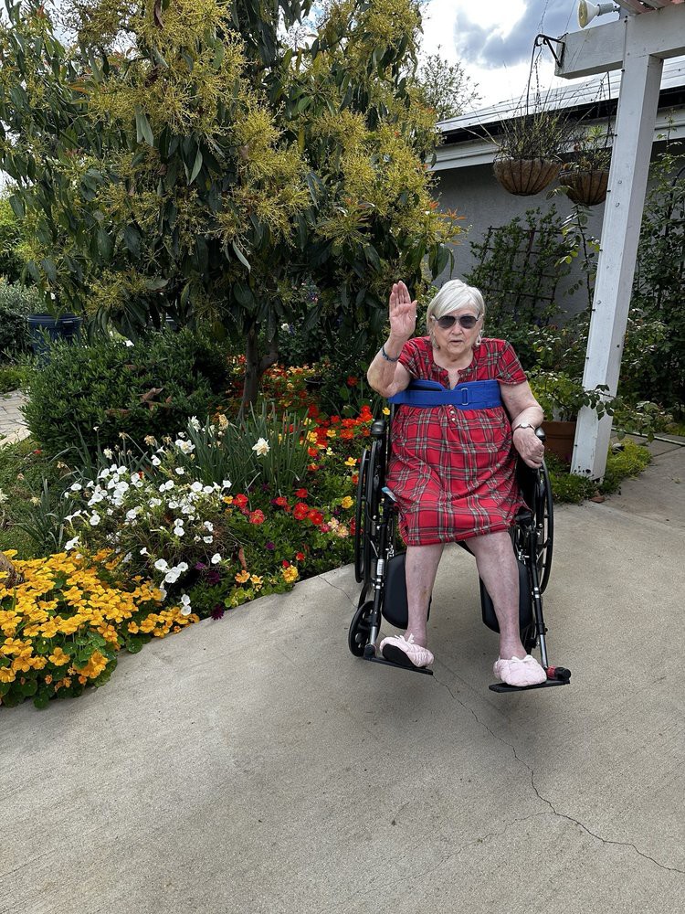 An elderly person in a wheelchair waves while sitting outdoors among colorful flowers.