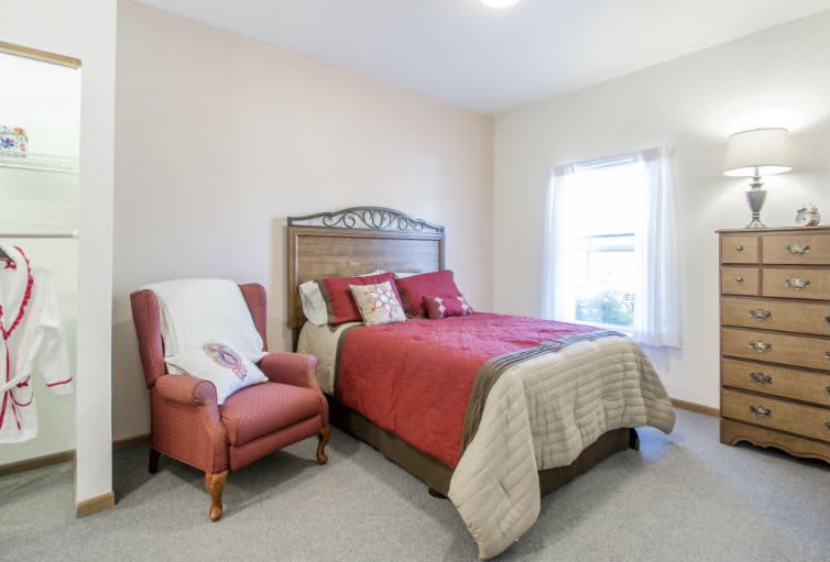 A cozy bedroom with red bedding and an armchair next to a window.