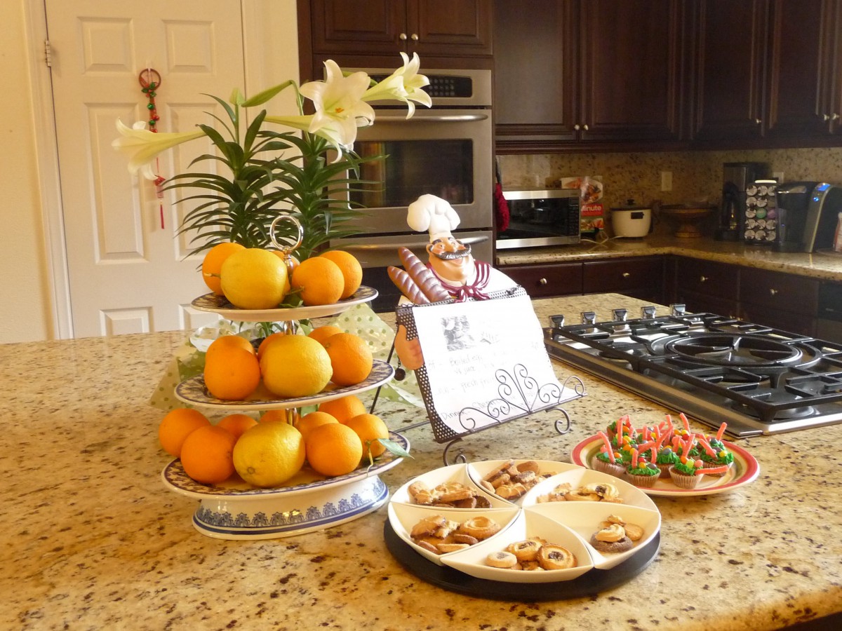 A kitchen scene with a tiered fruit stand of oranges, decorated cookies on plates, and a luxurious granite island countertop.