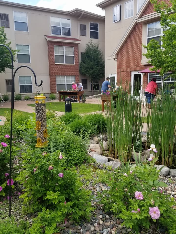 A beautifully maintained garden area with lush greenery and a small pond, set against the backdrop of an apartment complex. People are enjoying the serene environment.
